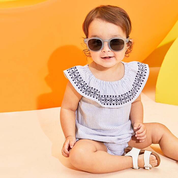 A female model in sunglasses, white sandals, and a summer onesie.
