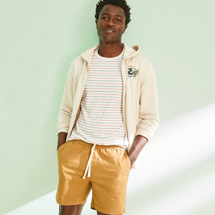 A model wears a cream colored zip-up hoodie over a striped t-shirt and twill drawstring jogger shorts