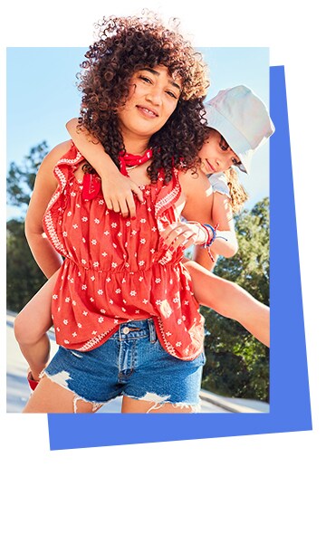 A female model wearing dark wash denim shorts and a short sleeve red top. A younger model on piggy back wearing a beige bucket hat.