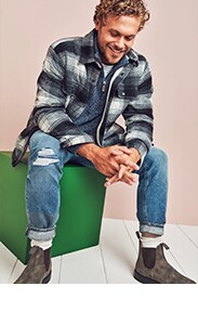 A male model wearing a blue flannel layered with a blue quarter zip, rolled jeans, and browl ankle boots.