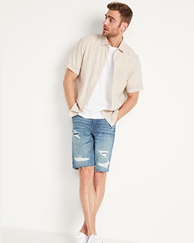 A male model wears a cream colored short sleeve button-up shirt & distress washed denim shorts.