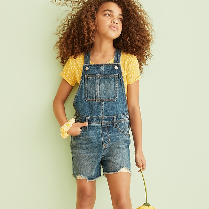 A female model with curly hair dressed in a dark denim shorts overall with a yellow top and yellow scrunchie on her wrist.