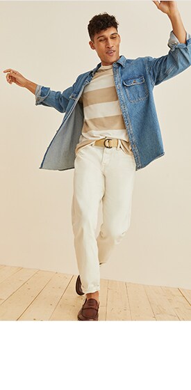 A male model wears a denim shirt over a cream and white horizontal striped t-shirt and cream colored jeans.