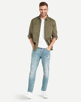 A male model wears Relaxed Slim Taper style denim and a green button up shirt over a white t-shirt.