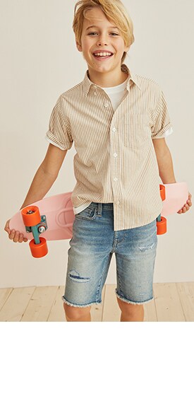 A boy wearing a striped buttondown, jean cutoff shorts, and holding a skateboard.