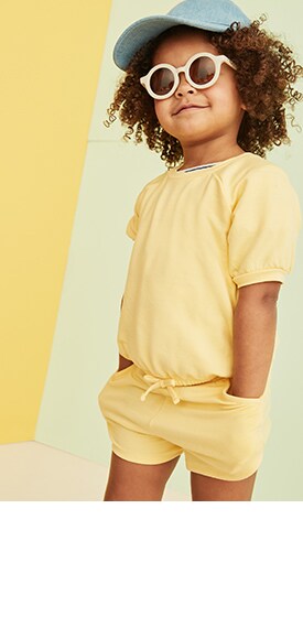 A model with curly hair, wearing whote round sunglasses, a baseball cap, and a soft yellow shorts romper.