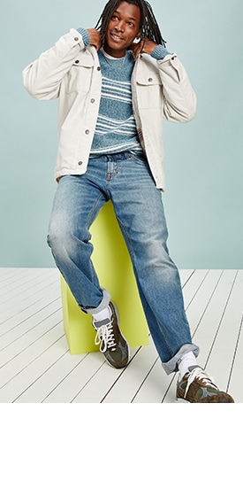A male model wears medium tint Boot-Cut Built-In Flex jeans, light colored barn-style jacket and striped sweater