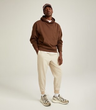 A male model wears activewear style pantss, & a light brown hoodie.