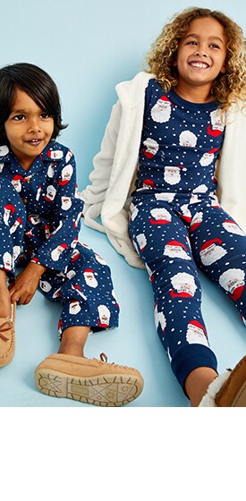 Young models sitting down and wearing Old Navy Santa Pajamas and slippers.