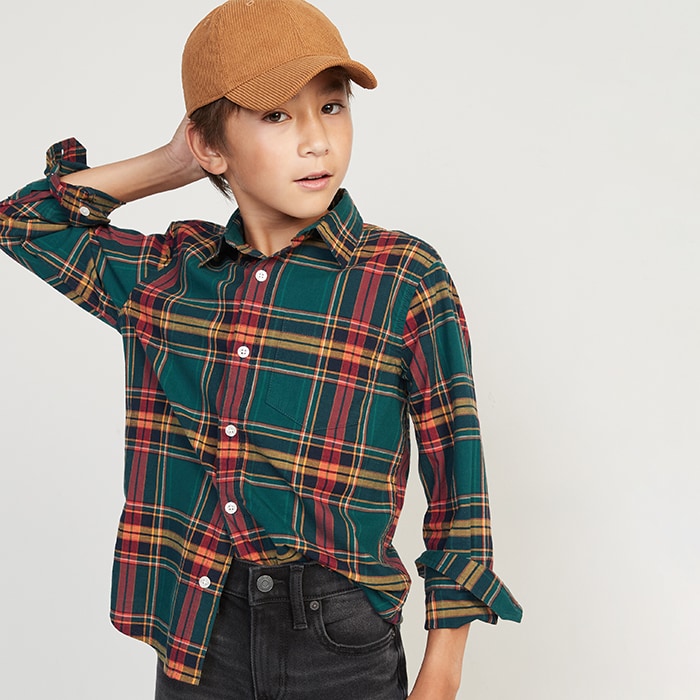 A young boy dressed in a green and red longsleeve flannel, dark black jeans, and a baseball cap.