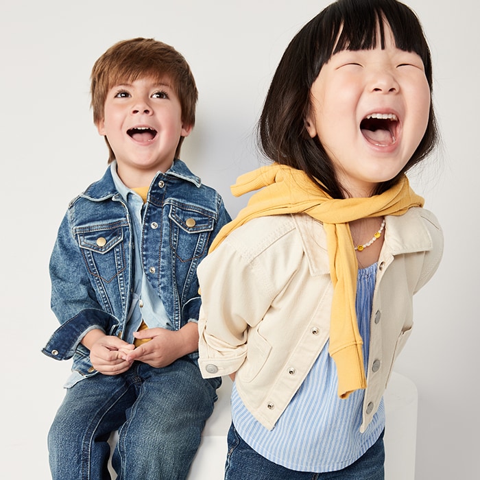 Two young models wearing blue jean jacket and jeans for toddler boys and blue striped top and beige jeans jacket for toddler girls.