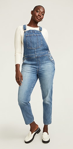 A model in a longsleeve cream top paired with light wash distressed overalls.