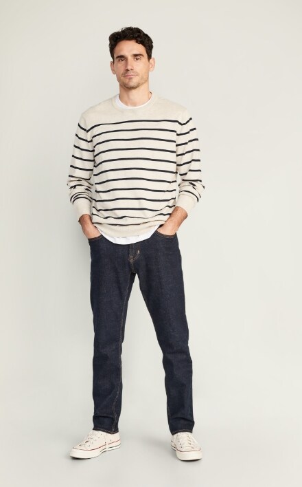 A male model wears dark washed Slim style jeans & a cream colored horizontal stiped sweater.