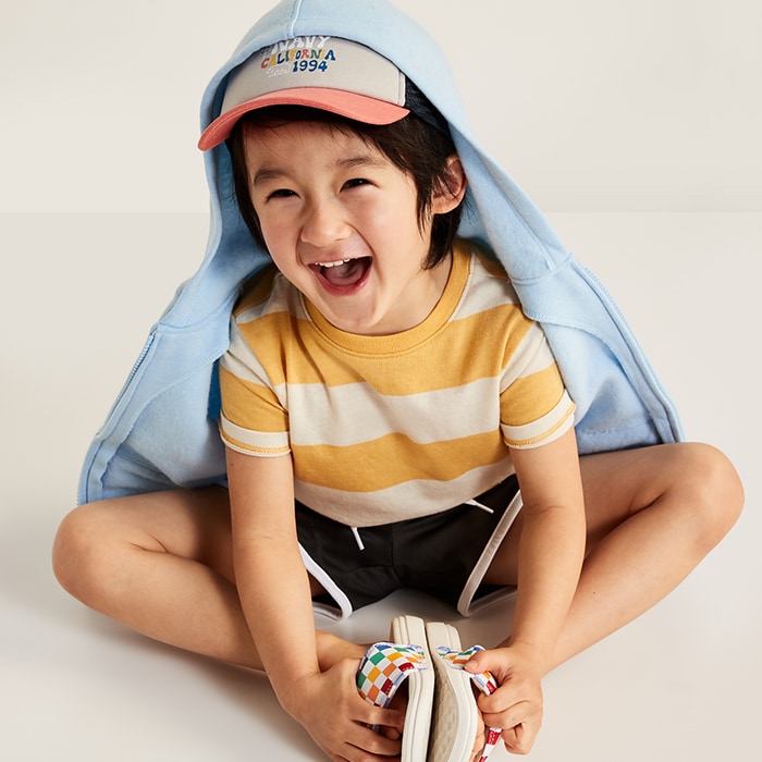 Young model wearing yellow striped t-shirt with black shorts, light blue zip hoodie slide sandals and a baseball cap.