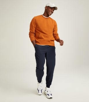 A male model wears an orange crew neck sweatshirt & dark activewear jogger bottoms.