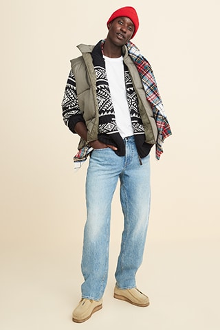 A male model dressed in light washed loose jean, holiday patterned sweater, flannel scarf and a red beanie.